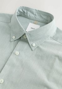 Hellgrünes Hemd mit Kragen aus strukturiertem Stoff, ausgestattet mit weißen Knöpfen und einem Etikett mit der Aufschrift "NEXT". Nahaufnahme zeigt Kragen und Nahtdetails.