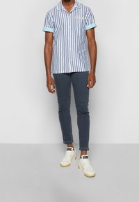 Kortgebreide, button-up shirt in blauw-witte strepen met lichtblauwe manchetten; gecombineerd met donkere jeans en witte sneakers.