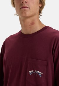 Mørkerød bomulds T-shirt med korte ærmer, rund halsudskæring og en brystlomme, der har et "Billabong" logo i blå og hvid.