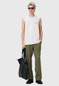 Off-white mouwloze shirt met tekst, olive groene cargo broek, zwarte leren tas en zwarte laarzen. Model heeft kort licht haar en een zonnebril.