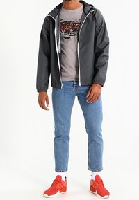 Schwarze Kapuzenjacke mit weißem Reißverschluss, graues Grafik-T-Shirt, hellblaue Jeans und rote Sportschuhe mit strukturierten Sohlen.