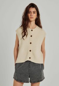 Stickad ärmlös tröja i ljus beige med ribbad textur och ett V-mönster. Har fem bruna knappar framtill. Matchas med grå shorts.