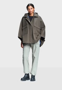 Femme portant une cape de pluie à capuche olive oversize, un pantalon cargo gris clair et des bottines noires, debout devant un fond uni.