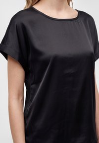 Femme portant un chemisier noir en satin à manches courtes avec un col rond et des manches retroussées, visible des épaules à la taille.