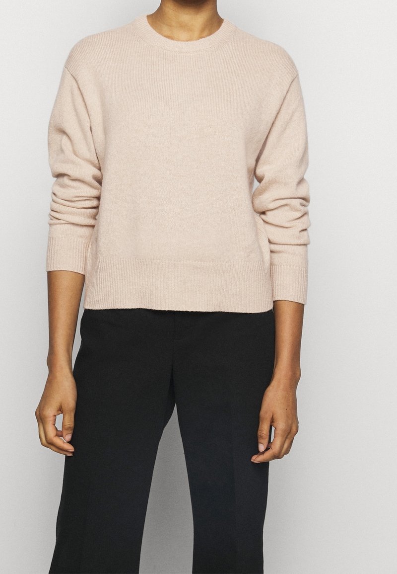 Personne portant un pull à manches longues rose pâle et un pantalon noir, debout devant un fond gris clair, les bras détendus.