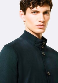 Giacca verde scuro con un colletto alto, caratterizzata da una texture liscia e quattro bottoni frontali. Il design è aderente, evidenziando un aspetto moderno.