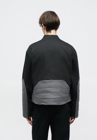 Casaco preto com painel traseiro e mangas acolchoados em cinza. Apresenta um colarinho, fecho de correr e um corte descontraído. Tecido de textura suave.