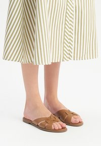 Sandali in pelle marrone a fascia incrociata con suola piatta, abbinati a una gonna a righe di lunghezza kneee in oliva e bianco.