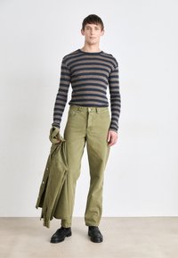 Chemise à rayures à manches longues en bleu marine et marron, pantalon vert olive, tenant une veste assortie. Chaussures noires à bout rond avec un détail texturé.