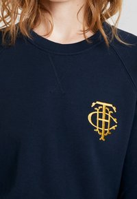 Marinblå sweatshirt med rund halsringning och raglanärmar; har en guldbroderad logotyp på bröstet. Tyget verkar mjukt och texturerat.