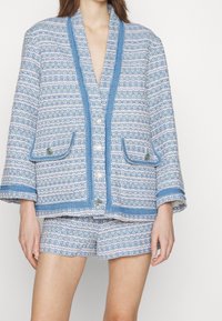 Ljust blå strukturerad jacka med mönstrad tyg, kontrasterande blå kant och knappar i guldton, ihopparad med matchande shorts.