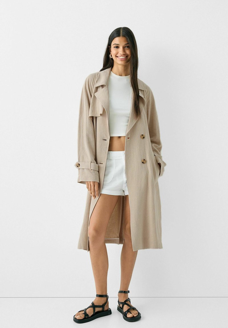 Trench coat di colore beige chiaro con una vestibilità rilassata, design doppiopetto e spacchi laterali. Abbinato a un top corto bianco, pantaloni shorts in denim e sandali neri.