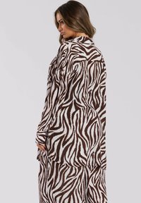 Camisa de estampado de cebra marrón y blanca con mangas largas, un corte relajado y un dobladillo ligeramente curvado, que muestra una tela texturizada.