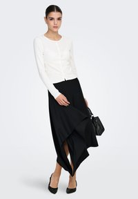 Cardigan blanc à manches longues avec boutons, associé à une jupe asymétrique noire à rayures subtiles, complétée par des talons noirs et un sac à main.