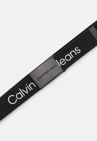 Zwarte stoffen riem met een rechthoekige metalen gesp waarop "Calvin Klein Jeans" is gegraveerd. Gladde textuur en gedurfde witte logo.