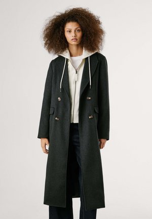 Cappotto in misto lana verde scuro con bottoni doppi, revers a punta e tasche. Indossato sopra un felpa bianco con zip.