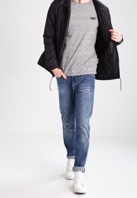 Giacca nera imbottita sopra una maglietta grigia melange, jeans in denim blu e sneakers bianche. Cuciture visibili e un piccolo logo sulla maglietta.