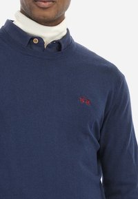 Mörkblå tröja med rund halsringning och ribbade ärmmuddar. Har en liten röd broderad logotyp på bröstet. Bärs över en vit polotröja.