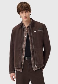 Brązowa kurtka z kordurowego materiału z zamykaną kieszenią, zestawiona z koszulą w kratę w odcieniach brązu, szarości i bieli. Czarne spodnie i pasek.