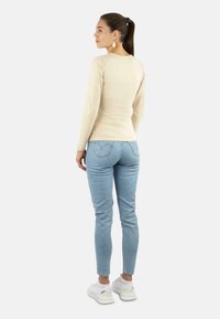 Beige Langarmoberteil, körperbetonter Stil; kombiniert mit hellblauen Skinny-Jeans; weiße Sneaker; Frau mit Pferdeschwanz und goldenen Ohrringen, von hinten betrachtet.