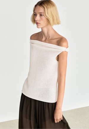Jeune femme aux cheveux blonds courts porte un haut en maille blanc épaules dénudées et un pantalon noir ample devant un fond clair uni.