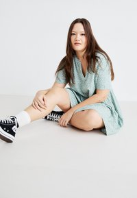 Green patterned dress with a V-neck and short sleeves, paired with black high-top sneakers and white crew socks, sitting on the floor.