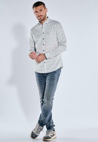 White button-up shirt with black floral pattern, paired with blue denim jeans and grey athletic shoes. Model stands upright with hands clasped.