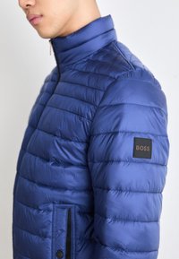 Doudoune bleue avec texture matelassée horizontale, col haut et un écusson carré noir portant l'inscription "BOSS" sur le bras supérieur.