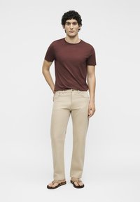 Homme debout portant un t-shirt bordeaux, un pantalon beige et des tongs beiges devant un fond clair uni.