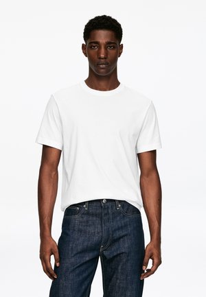 Jeune homme portant un t-shirt blanc uni à manches courtes et un jean bleu foncé, debout devant un fond blanc.