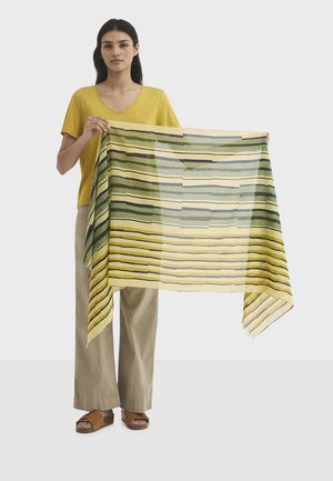 Femme portant un haut jaune et un pantalon beige tient une grande écharpe rayée jaune et verte étirée devant elle.