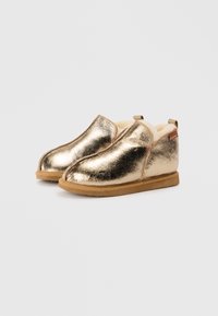 Chaussures slip-on métalliques dorées avec doublure blanche douce, bout arrondi et semelle en caoutchouc beige. Présentent une languette à l'arrière.
