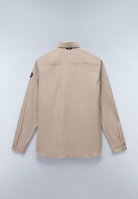Camisa de manga larga beige hecha de tela suave. Cuenta con un cuello con botones, puños de dos botones y un parche con el logo en la manga izquierda.