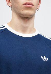 Námořnická modrá bavlněná mikina s bílým kulatým výstřihem, má tři bílé pruhy na ramenou a malé bílé logo na hrudi.