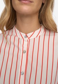 Skjorte med knapper, der har en hvid bund med lodrette røde striber, en rundet krave og blå knapper langs frontåbningen.