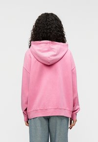 Ανοιχτό ρο ζ φούτερ με κουκούλα και κορδόνι, χαλαρή εφαρμογή, χαμηλωμένοι ώμοι και ριμπ μανσέτες, συνδυασμένο με ξεβαμμένο τζιν παντελόνι.