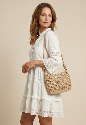 Femme en robe blanche en dentelle tenant un sac à bandoulière tissé couleur cuir clair sur un fond beige uni.