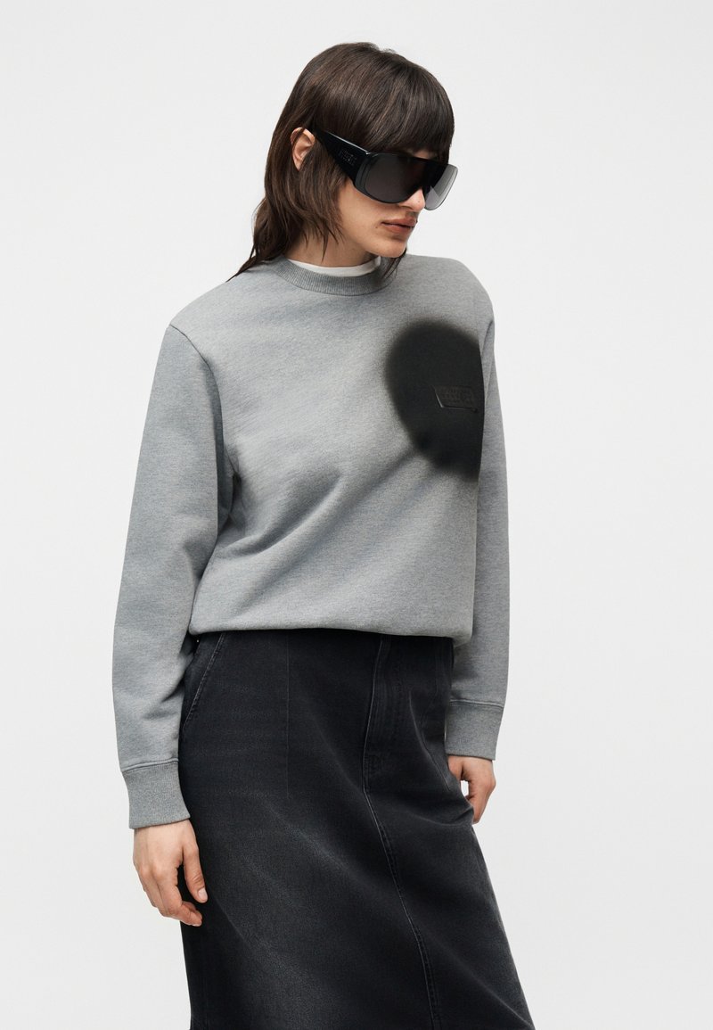 Femme portant des lunettes de soleil noires surdimensionnées, un sweat-shirt gris avec un imprimé circulaire noir, et une jupe noire, debout devant un fond uni.