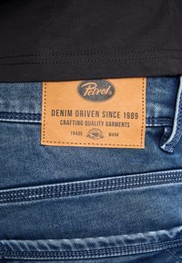 Džínové modré kalhoty s hnědou koženou nášivkou s logem "Petrol" a textem "DENIM DRIVEN SINCE 1989." Viditelné detaily šití.