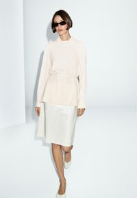 Blouse longue beige clair à manches longues avec une coupe décontractée et un col rond, associée à une jupe crayon argent métallisé arrivant au genou et des chaussures claires.