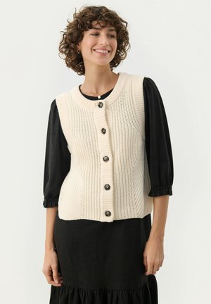 Gilet beige en tricot avec un motif texturé, encolure ronde et boutons noirs, porté par-dessus un haut à manches bouffantes noir.