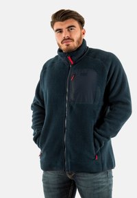 Giacca in pile blu navy con colletto alto, chiusura con zip e tasca sul petto; presenta dettagli rossi e un tessuto morbido e texturizzato.