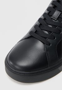 Μαύρο δερμάτινο αθλητικό παπούτσι με λείο και γυαλιστερό καπάκι, υφασμάτινες πλευρές και επίπεδα μαύρα κορδόνια πάνω σε δομημένη γλώσσα. Ελάχιστες λεπτομέρειες ραφής.