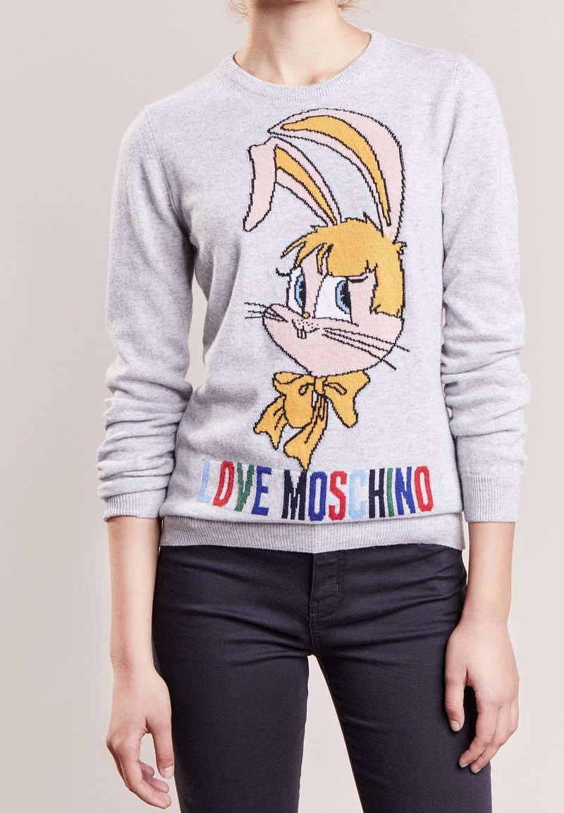 Personne portant un pull gris clair avec un visage de lapin dessiné et "LOVE MOSCHINO" en lettres colorées à l'ourlet, associé à un pantalon foncé.