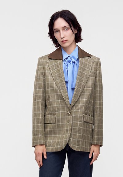 Jeune personne aux cheveux courts et foncés portant un blazer à carreaux beige avec col marron sur un chemisier bleu avec un nœud, debout devant un fond uni.