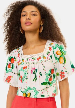 Frau mit lockigem Haar trägt große runde Ohrringe und eine weiße Bluse mit bunten Früchtemustern und korallenfarbene Hose.