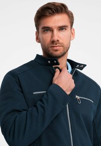Veste marine en tissu texturé, avec col montant et deux poches zippées. Présente des accents argentés et une coupe ajustée.
