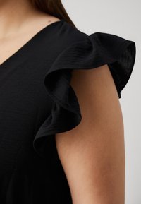 Närbild av en person som bär en svart ärmlös topp med volangdetaljer på axlarna mot en enfärgad bakgrund.