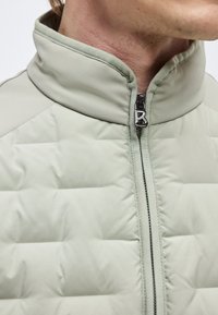 Άνδρας που φοράει ανοιχτό πράσινο επενδυμένο μπουφάν με ψηλό γιακά και εμπρόσθιο φερμουάρ που είναι μερικώς κλειστό.