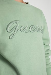 Felpa verde con logo "Guess" ricamato in rilievo dello stesso tono di verde. Presenta un classico girocollo e un tessuto morbido e strutturato.